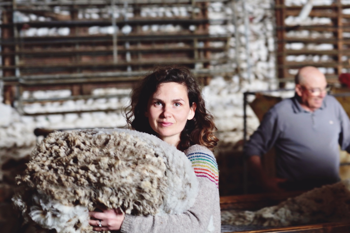 Solenn Couix-Loarer, a white woman with long curly brown hair wearing a gray wool sweater with rainbow stripes, holds a grayish-brown sheep fleece.