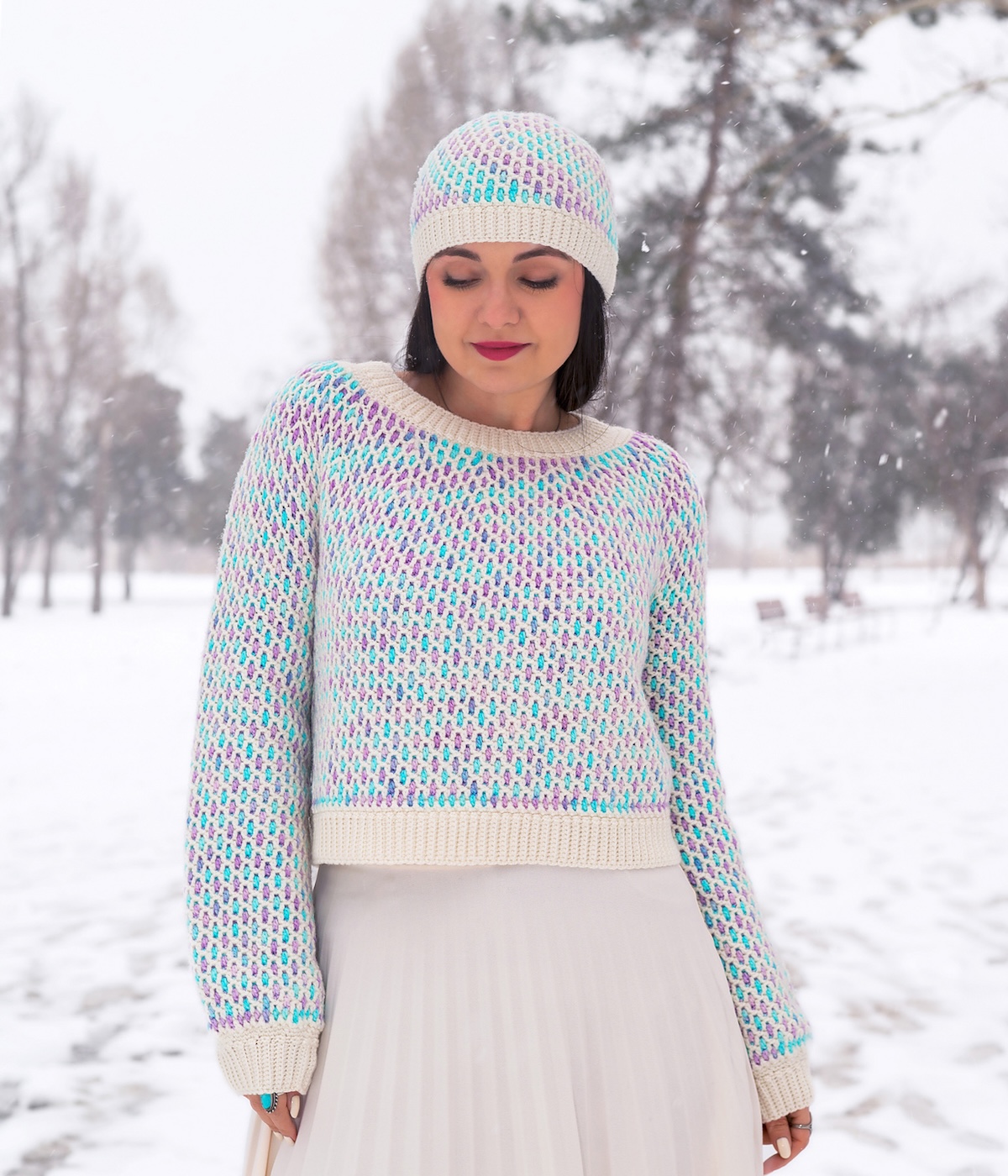 A light-skinned woman with dark hair stands in a snowy field modeling a crocheted beanie and sweater in frosty shades of white, blues, and purples.