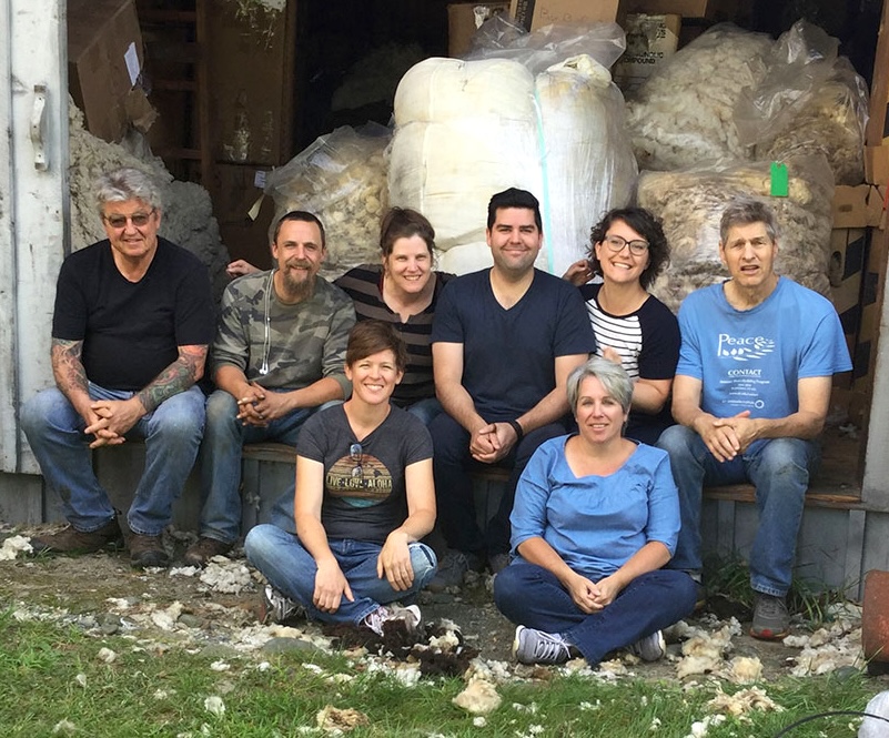 Eight members of the Julie Asselin Yarns team sit in front of a warehouse full of bales and bags of wool.