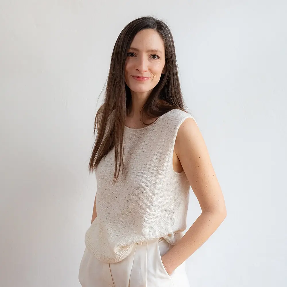 A light-skinned woman with long dark brown hair models a sleeveless knitted top in fuzzy white cotton yarn