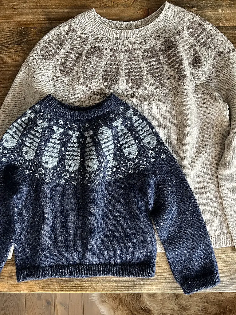 Two knitted sweaters are displayed on a wooden surface; one beige with a light brown and one dark blue with light blue, both featuring fish designs.