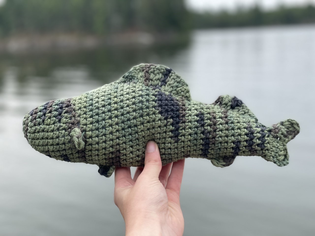 A person holds a green and gray crocheted walleye toy against a backdrop of calm water and trees.