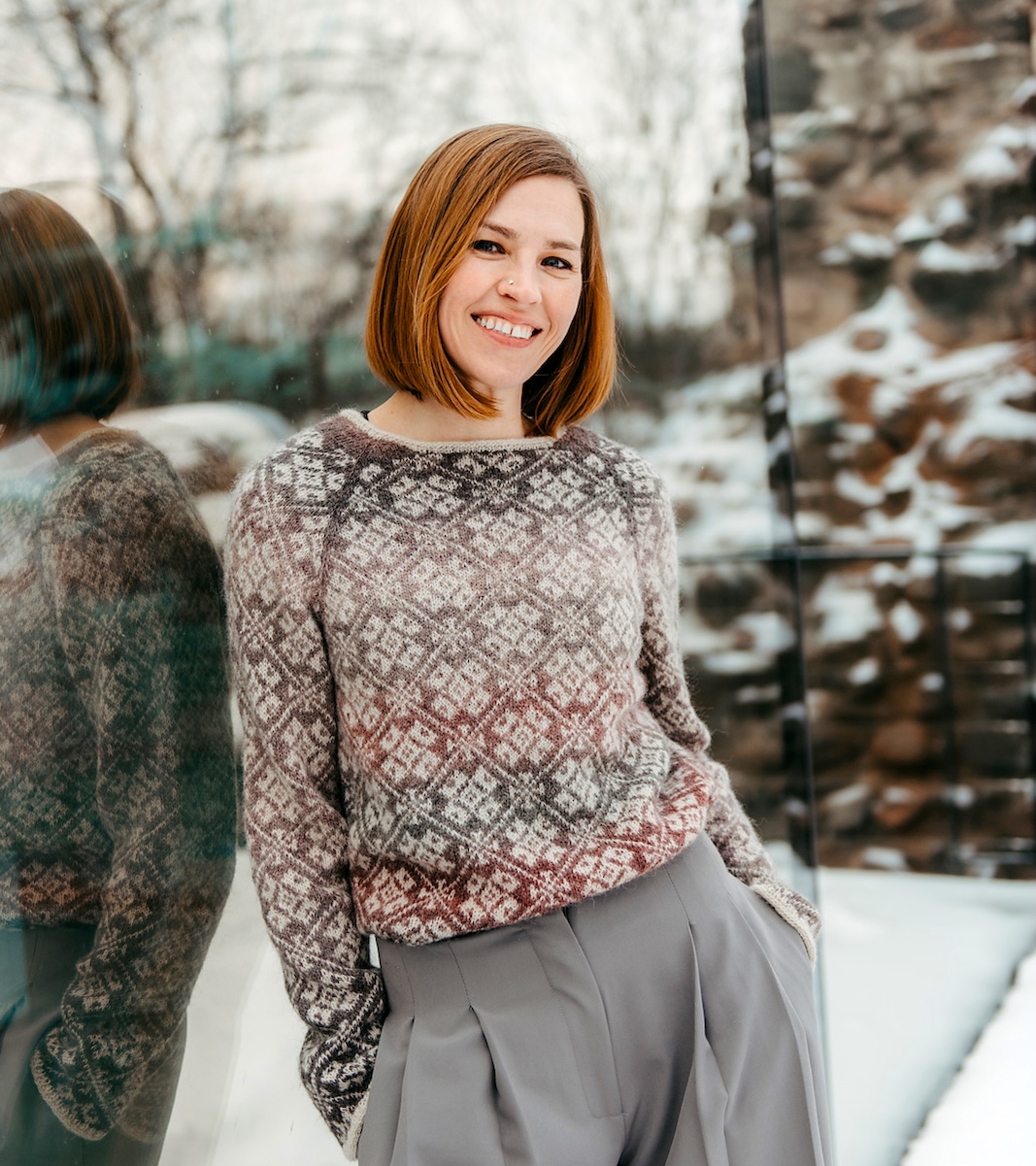A smiling woman with short red hair models a colorwork pullover knitted in white with the background in a color gradient yarn.