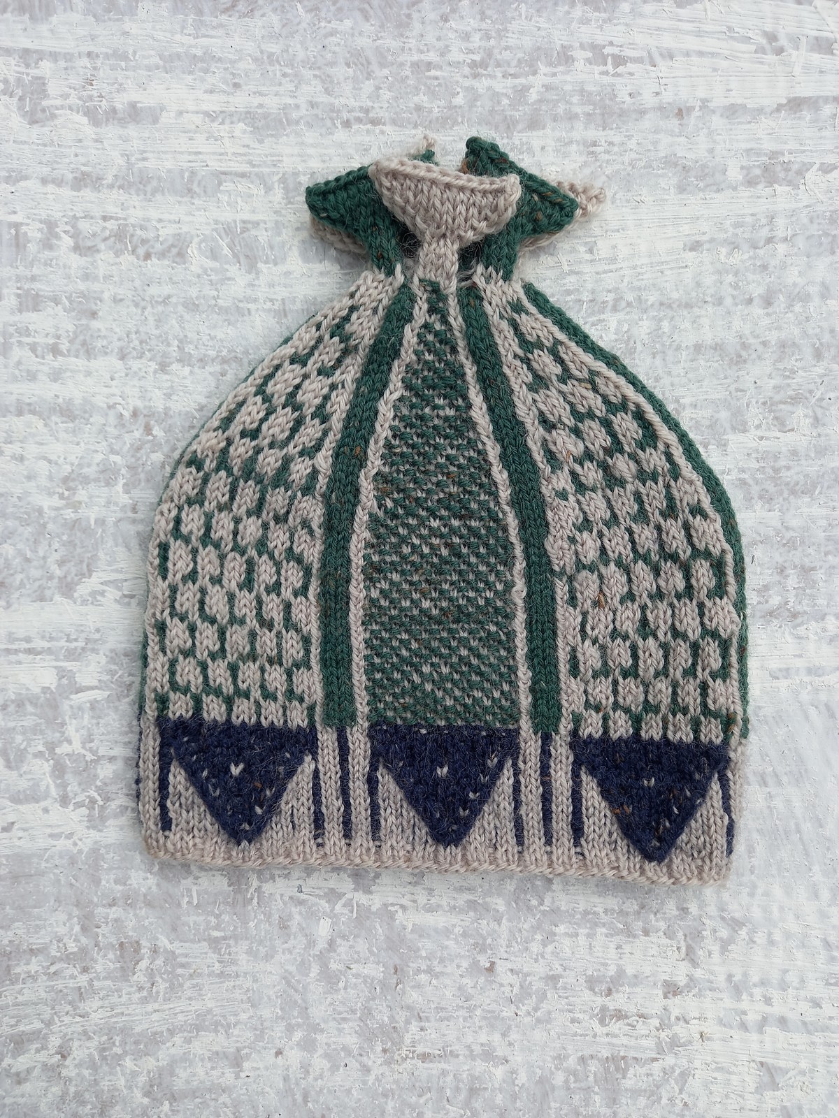 Knit hat with a geometric fish design in green, beige, and navy, featuring a gathered top and a textured pattern throughout.