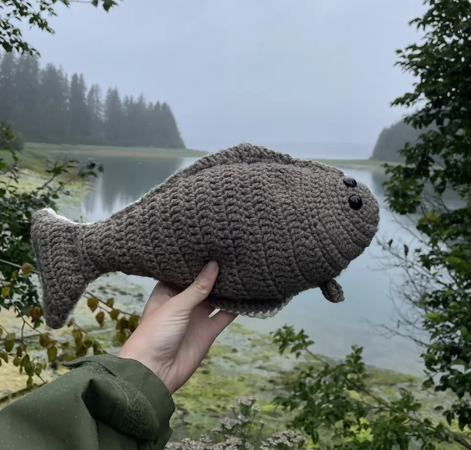 A person holds a crocheted gray halibut toy against a backdrop of a misty lake and evergreen trees.