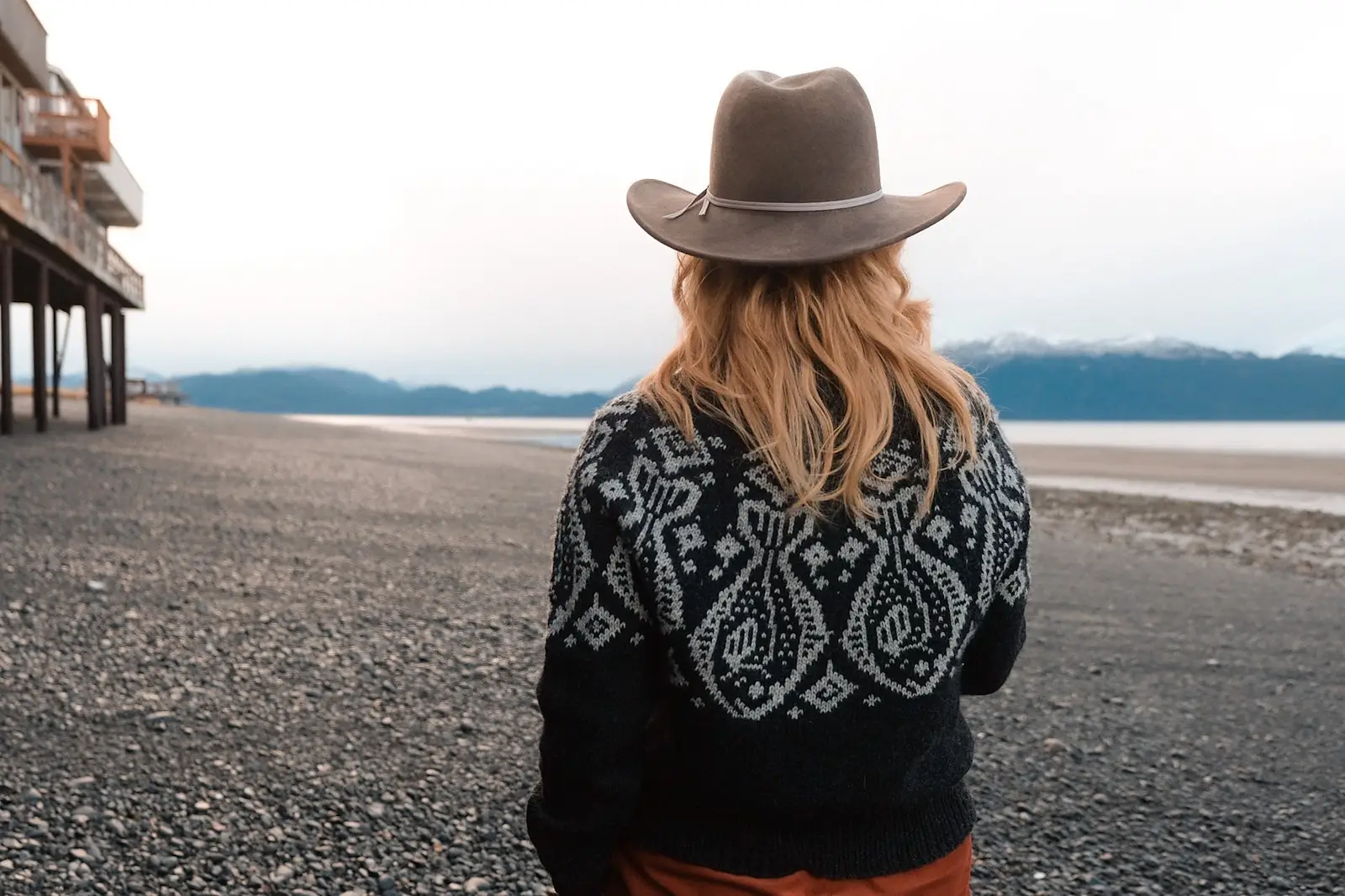 A person in a sweater featuring fish along the yoke stands on a rocky beach, gazing at the scenic mountains and calm water in the distance.