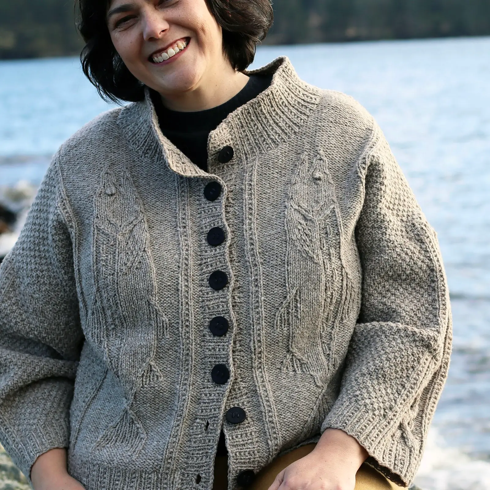 A person wearing a cozy, knit cardigan with fish patterns stands by a calm body of water, surrounded by nature.