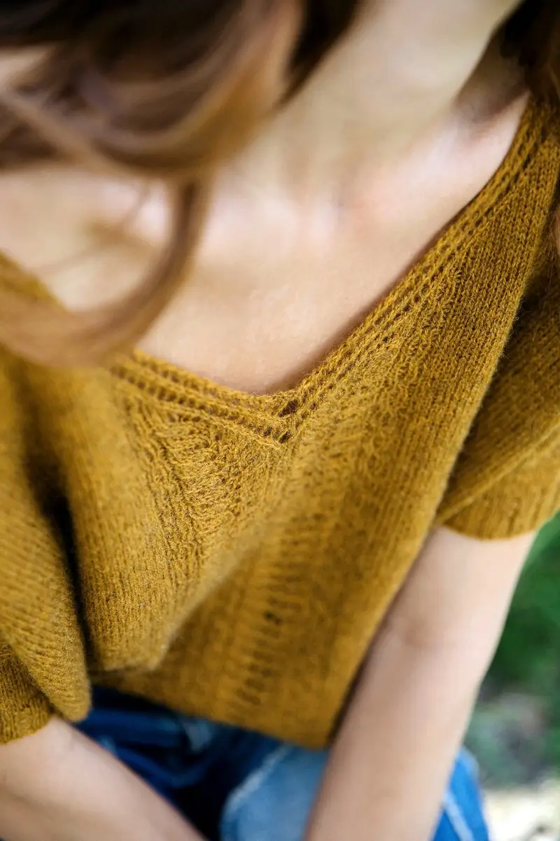 An extreme close-up of a person wearing a textured, mustard-yellow knit top with a deep V-neckline and short sleeves, paired with blue denim. The focus is on the intricate vertical eyelet and lace stitch patterns.