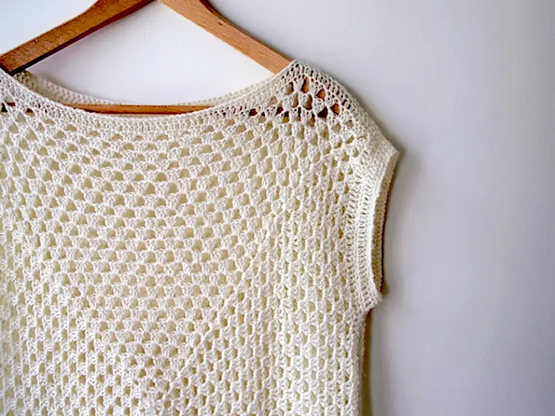 A close-up of the Amma Granny Square Top, a white, delicate open-knit crochet shirt with a classic mesh-like granny square pattern. It is displayed on a wooden hanger against a plain white wall, showcasing its sleeveless, boat-neck design and lightweight lace-weight texture.