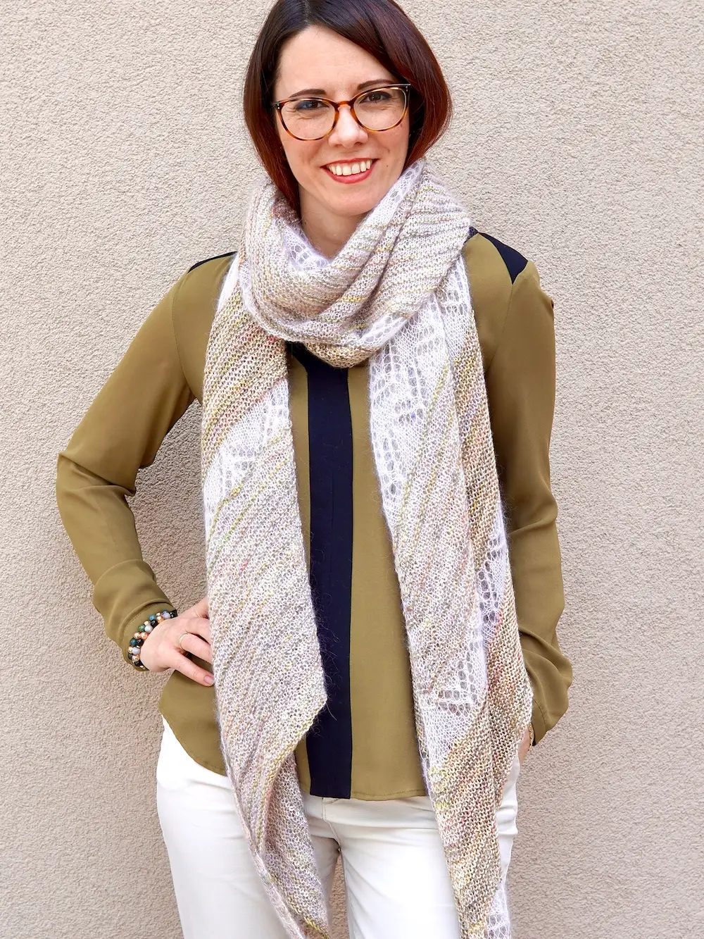 A white woman with short brown hair and glasses smiles at the camera. She is wearing an olive-green button-down shirt with a dark blue stripe down the center, white pants, and a lacy light-colored knitted shawl draped around her neck.