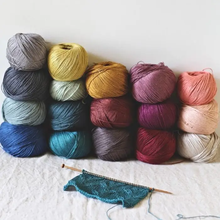 Several balls of silk yarn in assorted colors are stacked on a linen-covered table against a white wall, with a knitted lace swatch in deep teal lying in front of them.