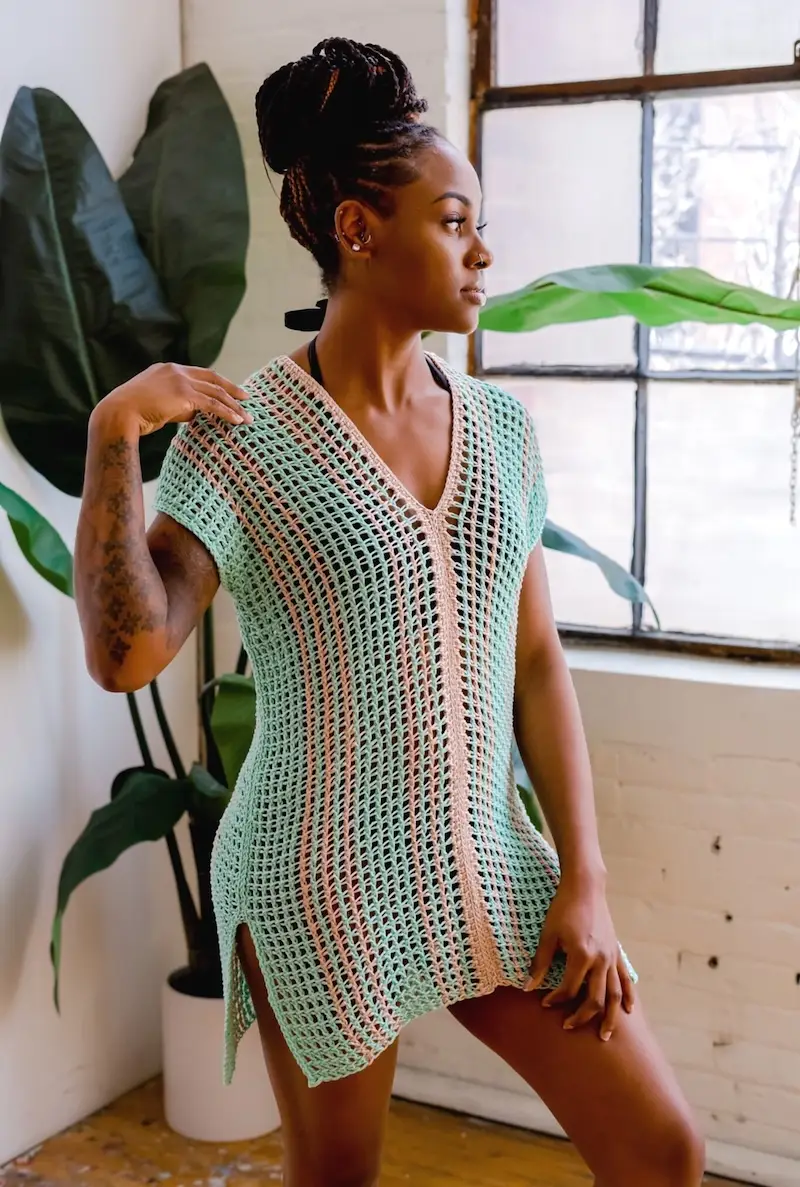 A person models the Aruba Tunic, a breezy Tunisian crochet top featuring a vertical mesh stitch pattern in mint green with pink stripes down the center and sides.