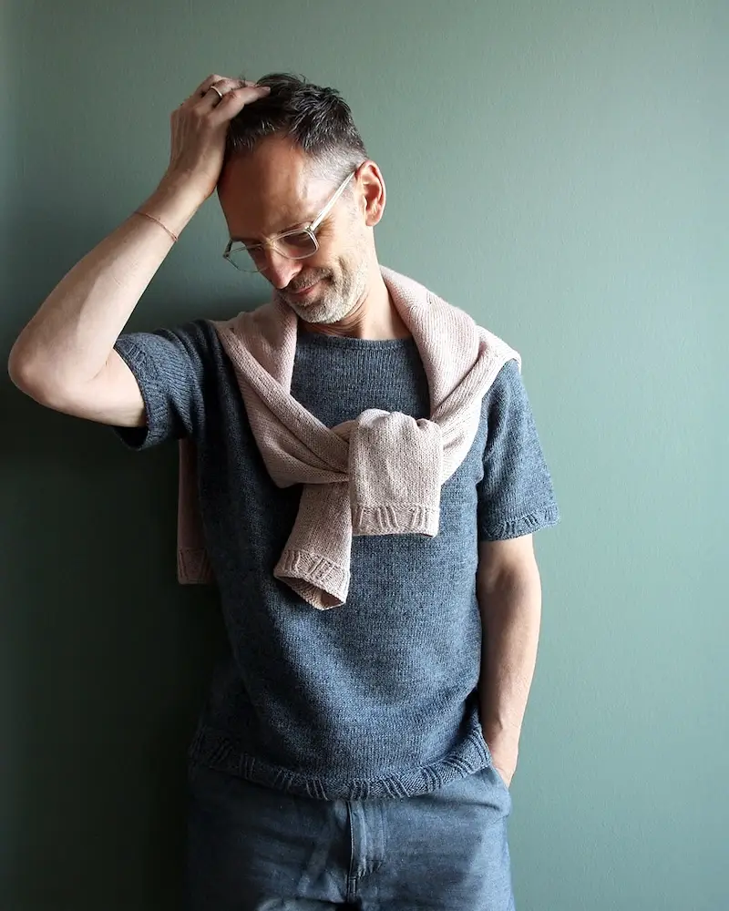 A man with glasses and short dark hair poses against a muted green wall. He is wearing a short-sleeved, charcoal grey knitted tee with a light pink sweater draped and knotted over his shoulders.