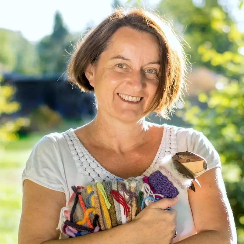 Britta Kremke, a middle-aged woman with brown bob-cut hair, smiles warmly at the camera. She is wearing a white scoop-neck t-shirt and standing outdoors in a sunlit garden. In her arms, she cradles a small, multi-colored sheep plush wrapped in various colors of knitted fabric.