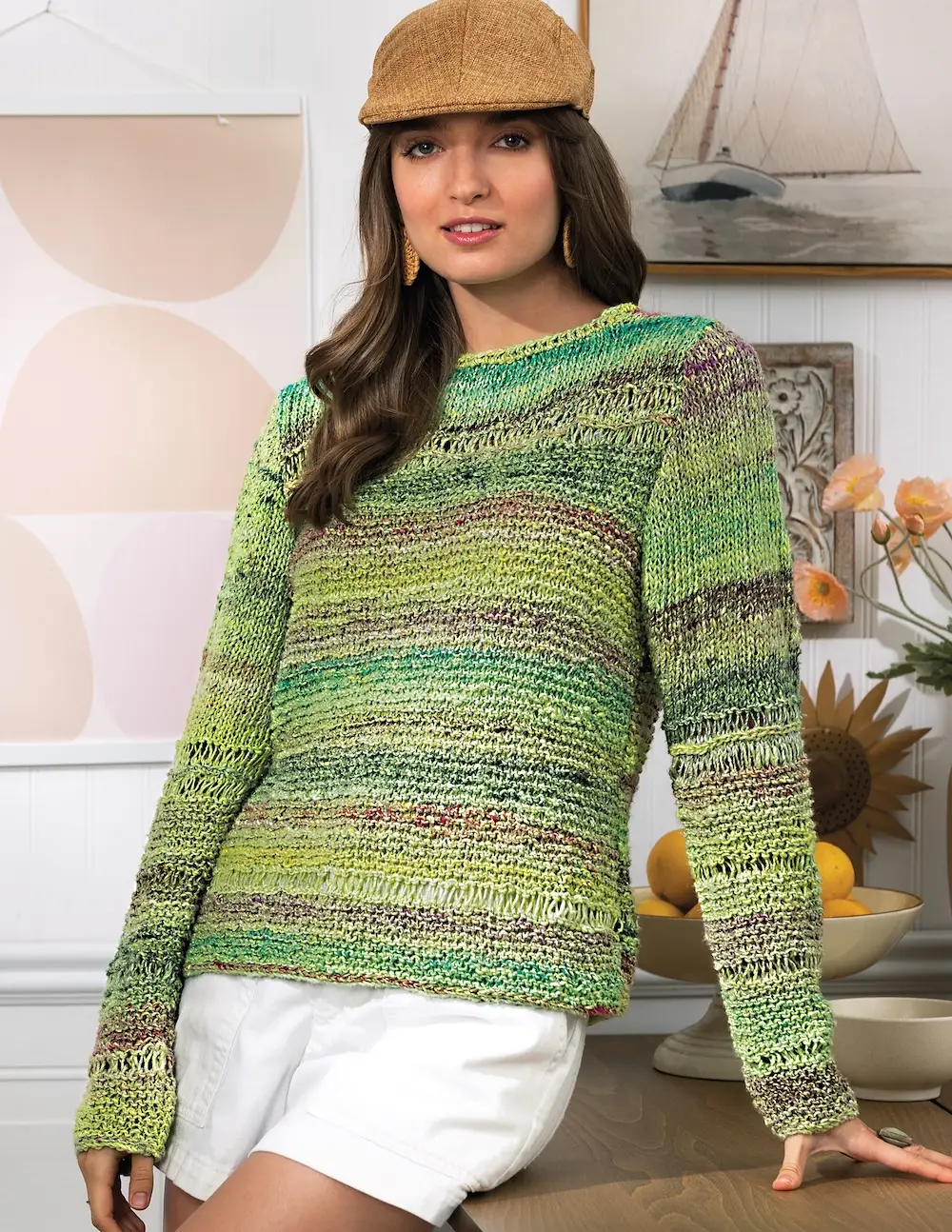A person with long brown hair wearing a tan flat cap and a textured, long-sleeved knit sweater. The sweater features horizontal, variegated stripes in shades of green, lime, and brown.