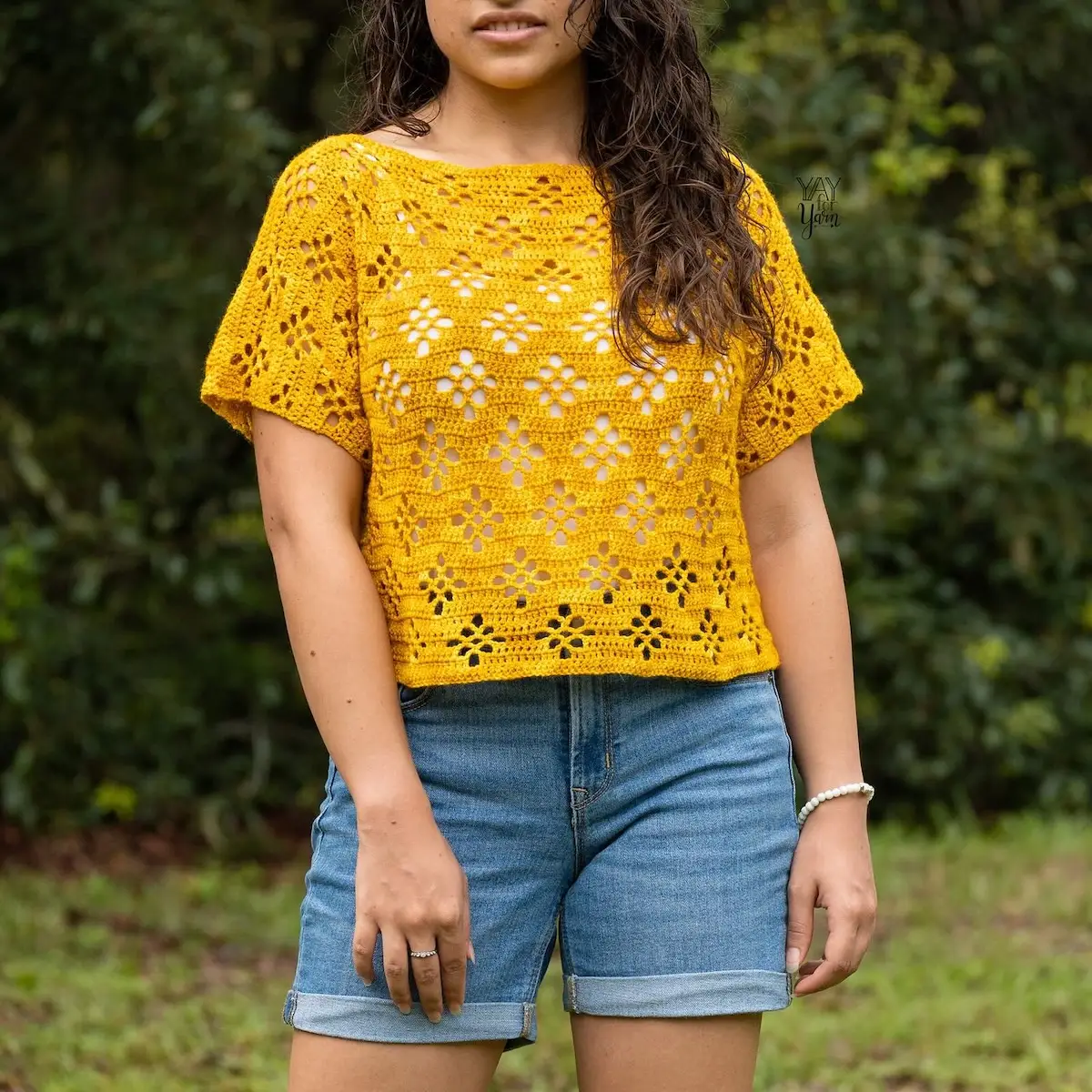 A person wearing a mustard yellow crochet lace short-sleeve top and denim shorts, standing outdoors against a soft green background.
