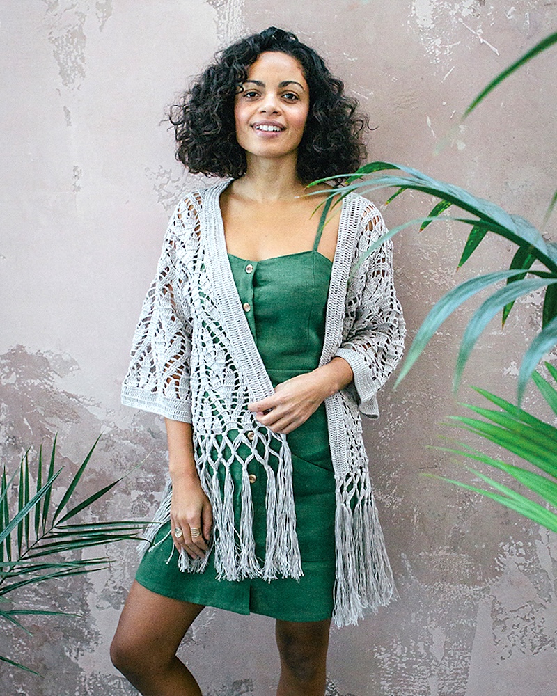 A smiling woman with dark, curly shoulder-length hair wearing a dark green, button-down sleeveless dress layered with a light grey, intricate crochet cardigan that features long fringe at the hem.