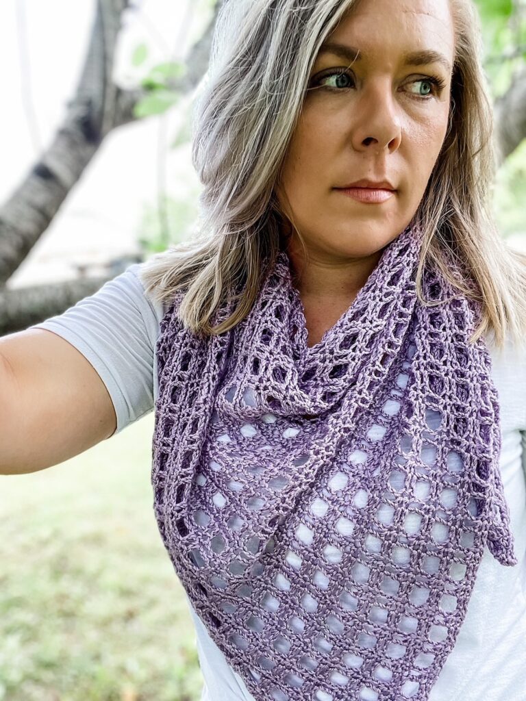 A woman in a white shirt outdoors wears a lavender crocheted scarf with a delicate lace pattern.