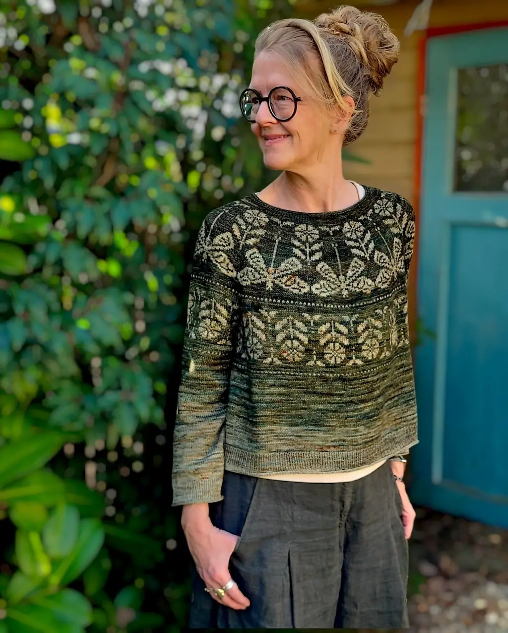 A smiling woman with glasses and her hair in a bun stands outdoors, wearing a knitted, long-sleeved sweater in dark green with an intricate, light-colored yoke. The design features a row of large dragonflies around the chest, framed by delicate floral and beetle motifs. She stands against a background of lush green foliage and a blue door.