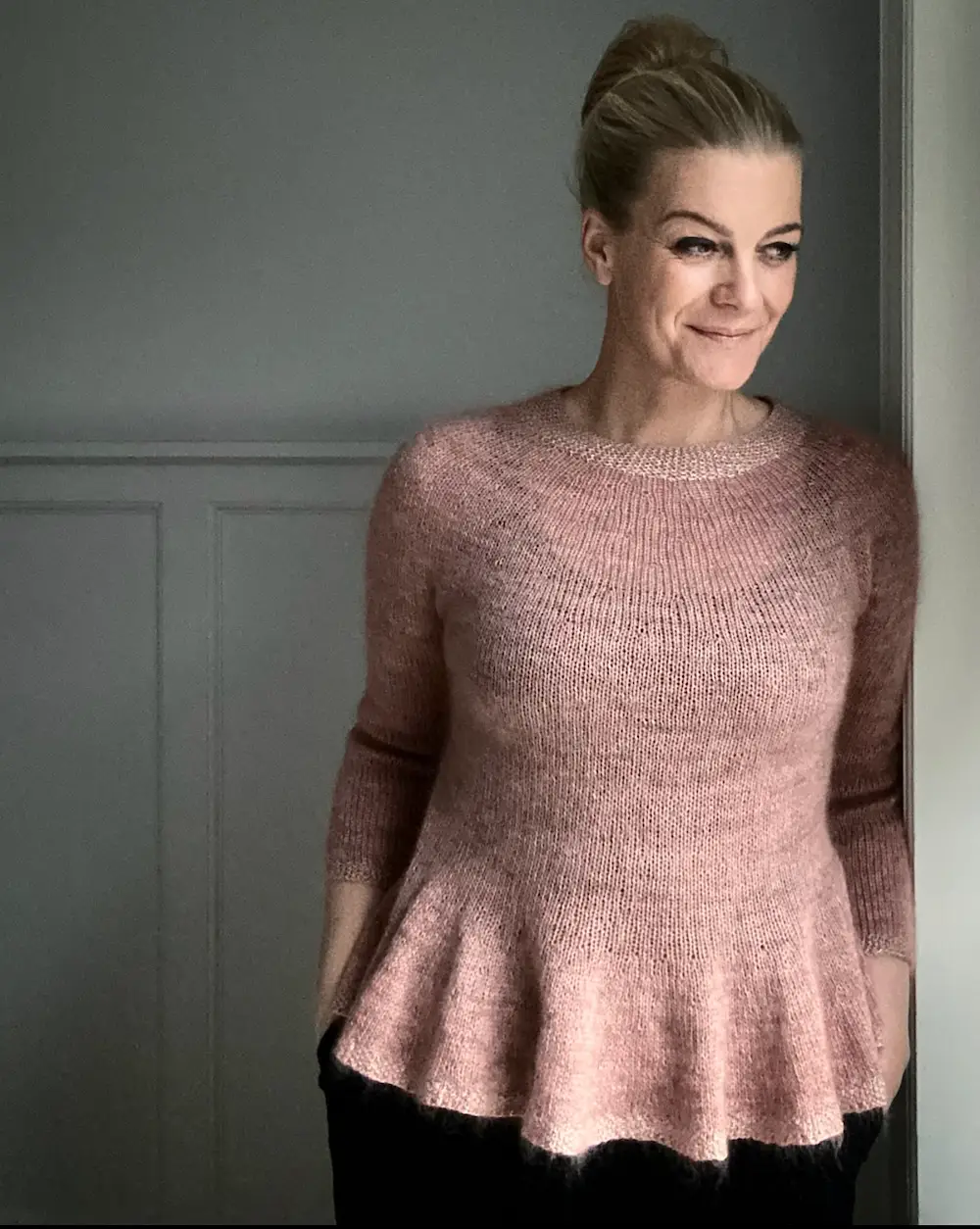A white woman with dark blonde hair pulled back stands against a gray paneled wall, wearing a dusty rose knitted peplum sweater. She leans against a white doorframe on the right, looking away from the camera with a slight smile. 