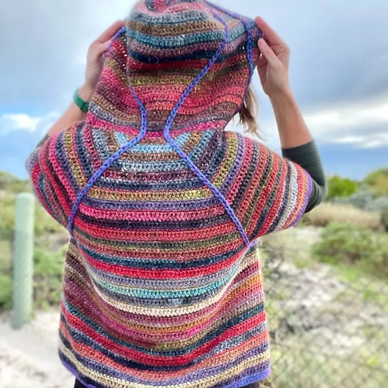 A person wearing a colorfully striped crochet hoodie stands with their back to us, looking out at sand dunes.