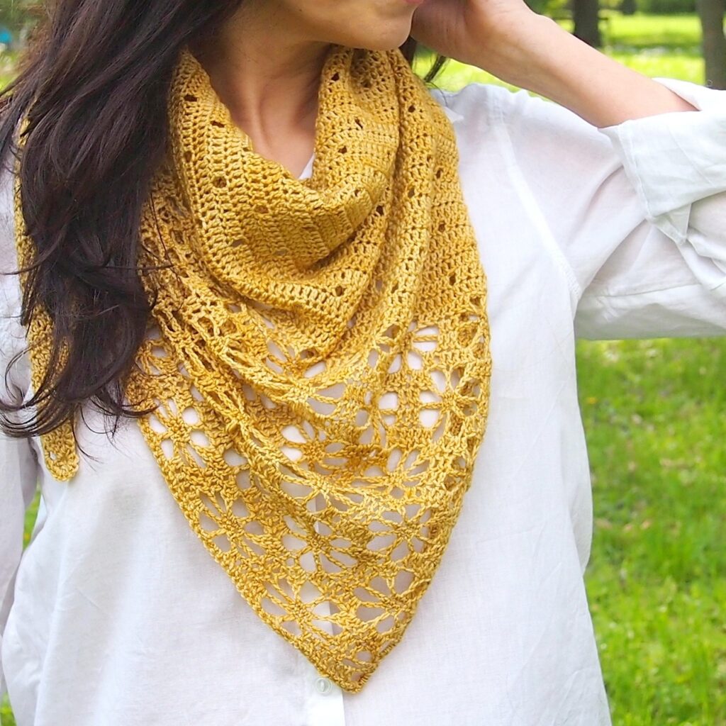 Woman wearing a white shirt and a yellow crocheted scarf with intricate patterns, standing outdoors against a blurred green background.