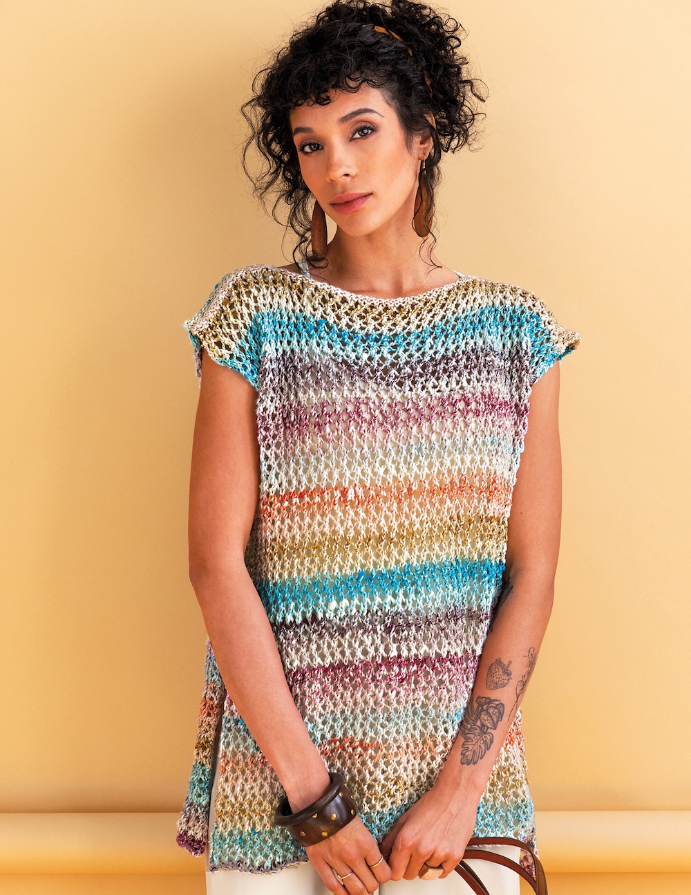 A woman with dark, curly hair tied up, wearing a colorful, open-knit, striped tunic with short sleeves and a boat neckline. The horizontal stripes feature a mix of blue, purple, orange, and white. 