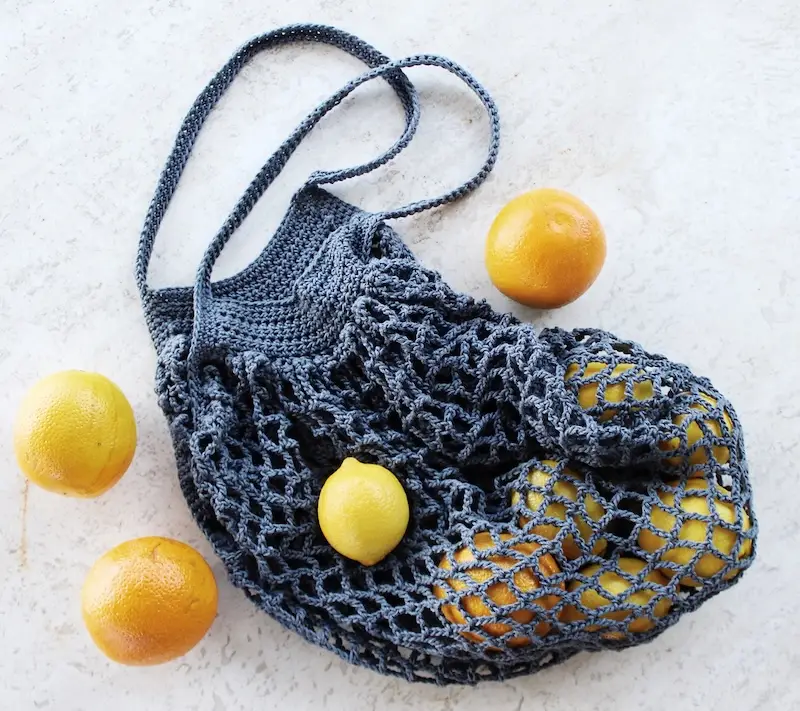A grey-blue crochet market bag resting on a light-colored, marbled surface. The bag features a solid, V-shaped top border with long shoulder straps and a wide-mesh, diamond-patterned net body. Several citrus fruits, including oranges and bright yellow lemons, are scattered around and nestled inside the bag.