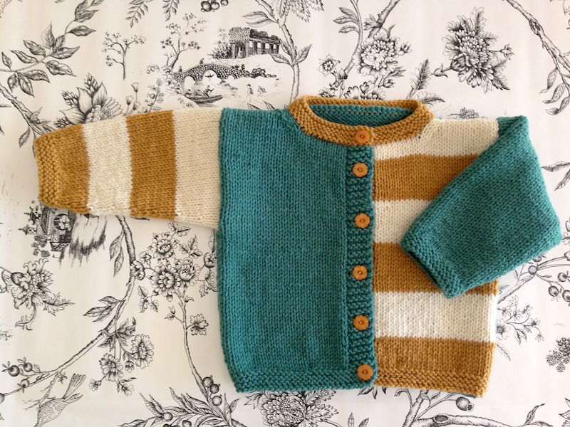 A hand-knitted baby cardigan with a playful, asymmetrical design. The left half of the sweater is solid teal, while the right half and sleeves feature bold horizontal stripes in cream and mustard yellow. The cardigan is finished with a mustard-yellow collar and a vertical row of five wooden buttons. It is laid against a white fabric background with a delicate black floral print.