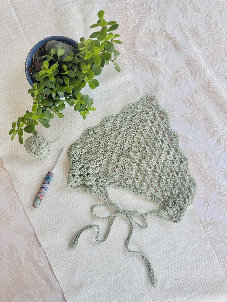 A delicate green crocheted kerchief with scalloped edges is laid on a textured white cloth beside a potted plant and crochet hook.