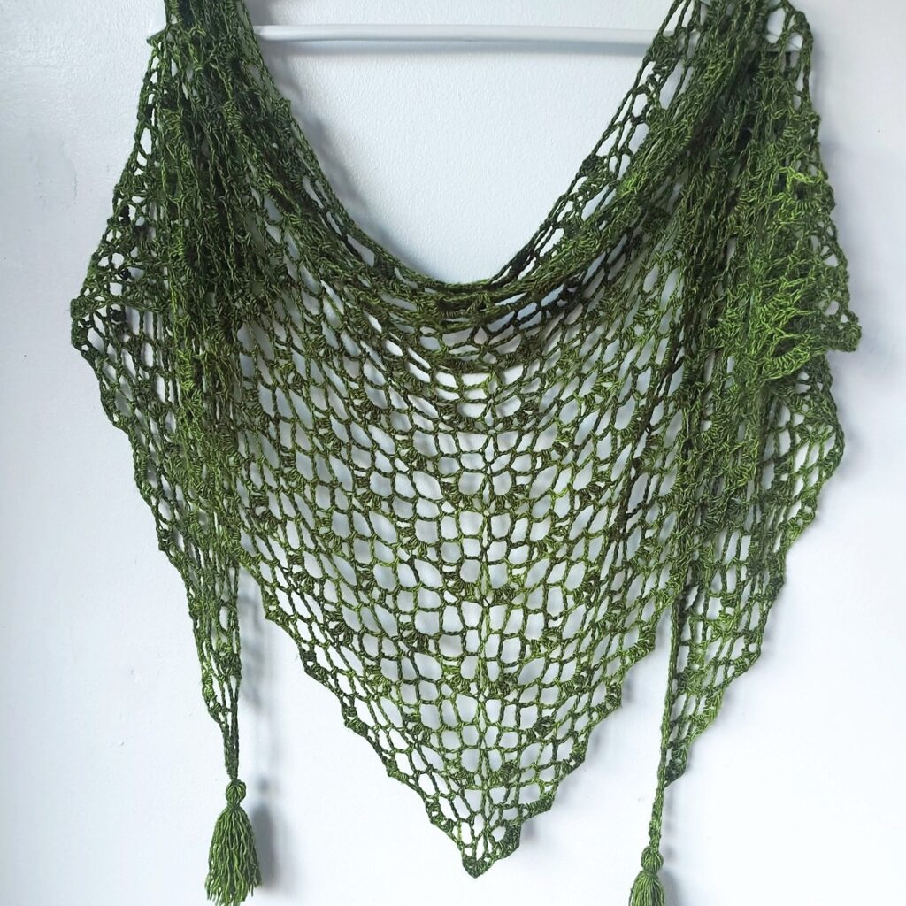 Green crocheted shawl with a triangular shape, featuring a delicate open stitch pattern and tassels, hanging on a white wall, evoking a rustic charm.