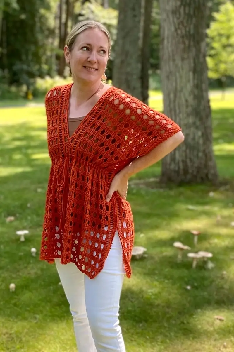 A woman standing outdoors in a grassy area with trees, modeling a burnt-orange crochet tunic. The tunic features a deep V-neckline, wide elbow-length sleeves, and a breezy, open-mesh stitch pattern. It has a high empire waistline and high side slits, reaching mid-thigh. She is wearing white pants underneath and has her left hand on her hip while looking off to the side with a smile.