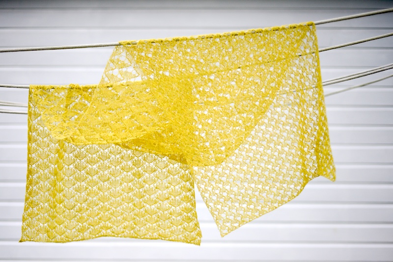 A yellow knitted lace shawl featuring a repeating geometric diamond pattern, hanging on a set of clotheslines. The intricate openwork design consists of interconnected diamond shapes with small lace eyelets creating a delicate, mesh-like texture.