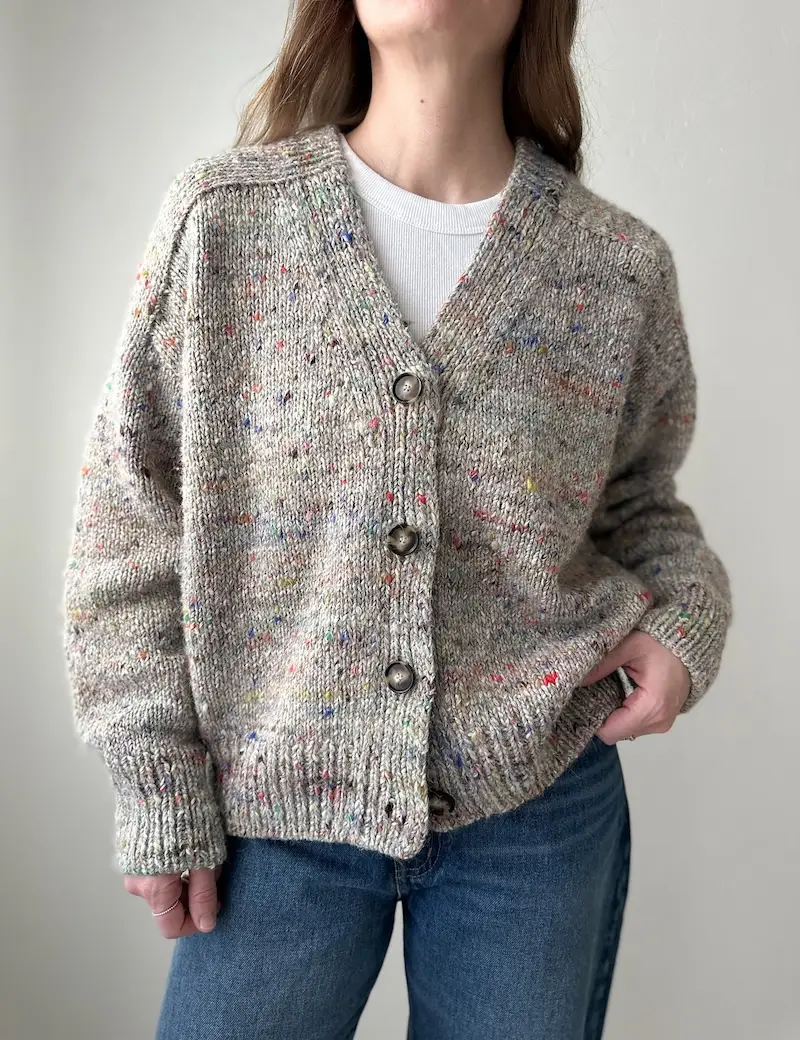 Closeup of a light-skinned person with long brown hair modeling a knitted V-neck cardigan in tweedy gray yarn with bright colored flecks.