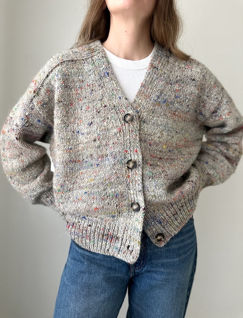 A person is shown from the neck down wearing the Lakes Cardigan by Ozetta, an oversized, V-neck, button-down sweater knit in a marled, speckled yarn.