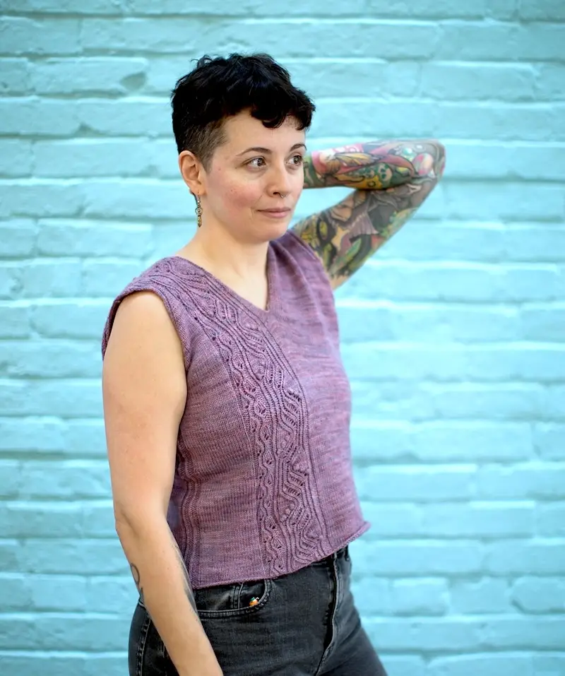 A light-skinned person with short, dark hair and visible tattoos on their left arm models a sleeveless, lilac-colored knit top with a V-neckline and a wide, vertical lace panel. The person stands in profile against a light turquoise brick wall, looking off-camera.
