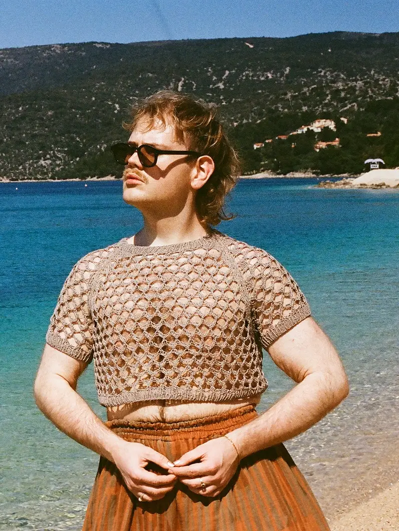 A person with light skin, short blonde hair, and a mustache stands on a pebble beach, looking to their left. They are wearing black sunglasses, a beige short-sleeved open-knit crop top, and a long, pleated brown skirt. The background features a vibrant turquoise and deep blue sea, with a large, green, tree-covered mountain rising under a clear blue sky.