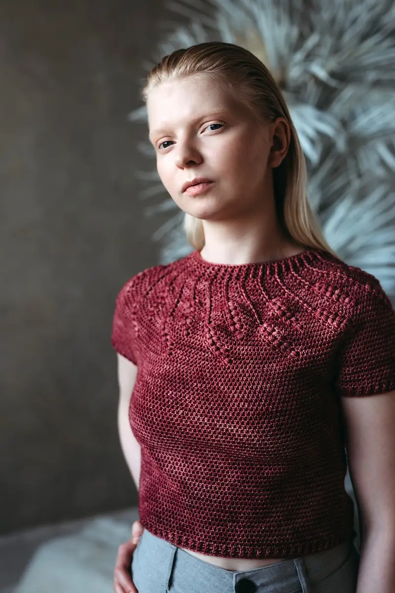 A woman with blonde hair wearing a short-sleeved, deep burgundy crocheted sweater with a textured circular yoke with raised leafy motifs. The background is a moody, dark grey with soft botanical shadows.
