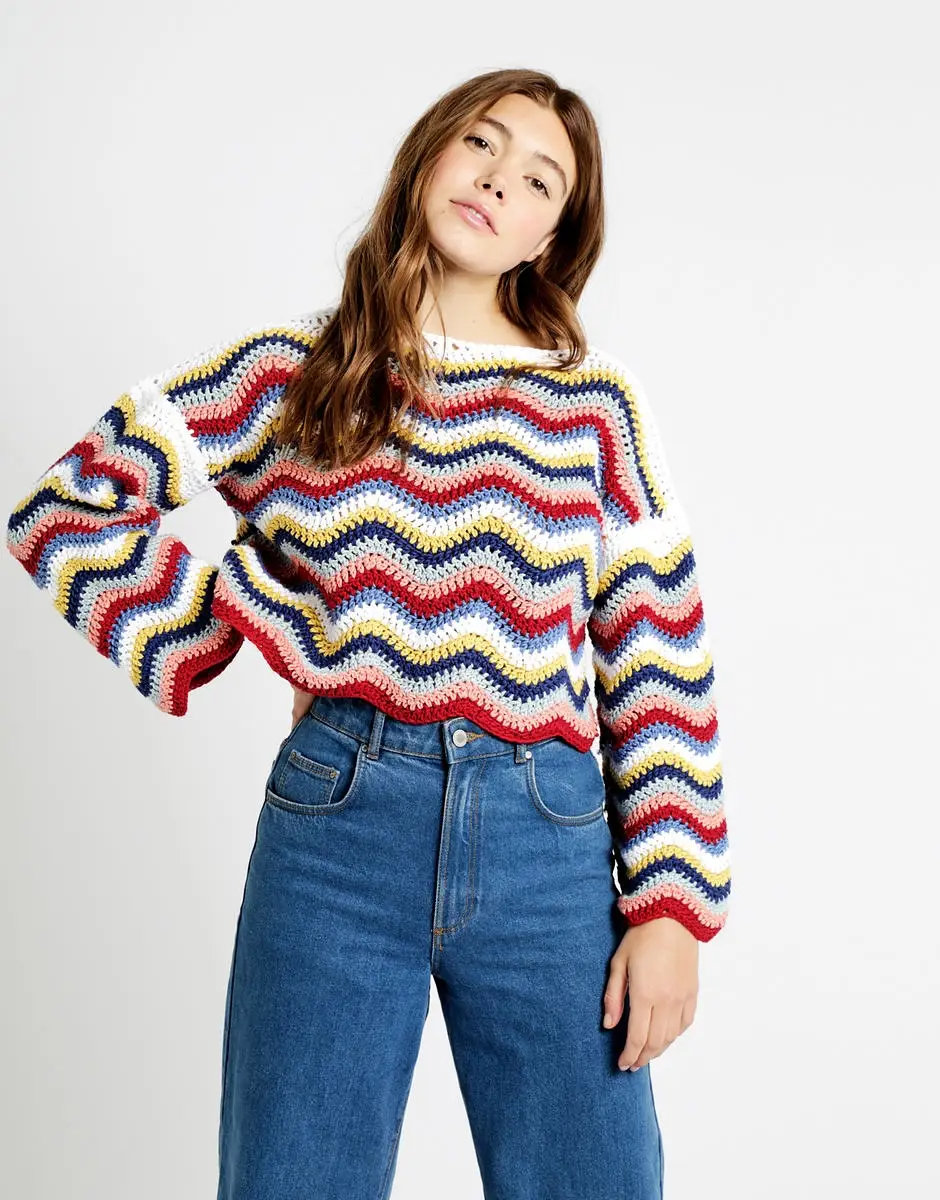 A young woman with long, wavy brown hair wears a vibrant, multi-colored crochet sweater in a bold chevron pattern. The sweater has a relaxed, slightly cropped fit with long sleeves. She pairs it with high-waisted, wide-leg blue jeans. She leans her head slightly to the side with a neutral expression.