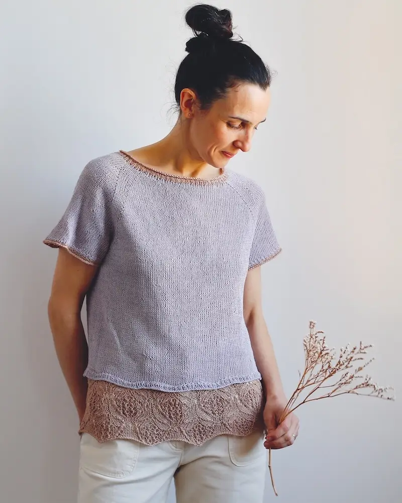 A woman with dark hair in a bun, wearing a short-sleeved, lavender-colored knitted top with a relaxed fit. The top has a simple crew neckline and subtle mauve-pink trim along the collar and sleeve edges, with a lace underlayer at the hem.