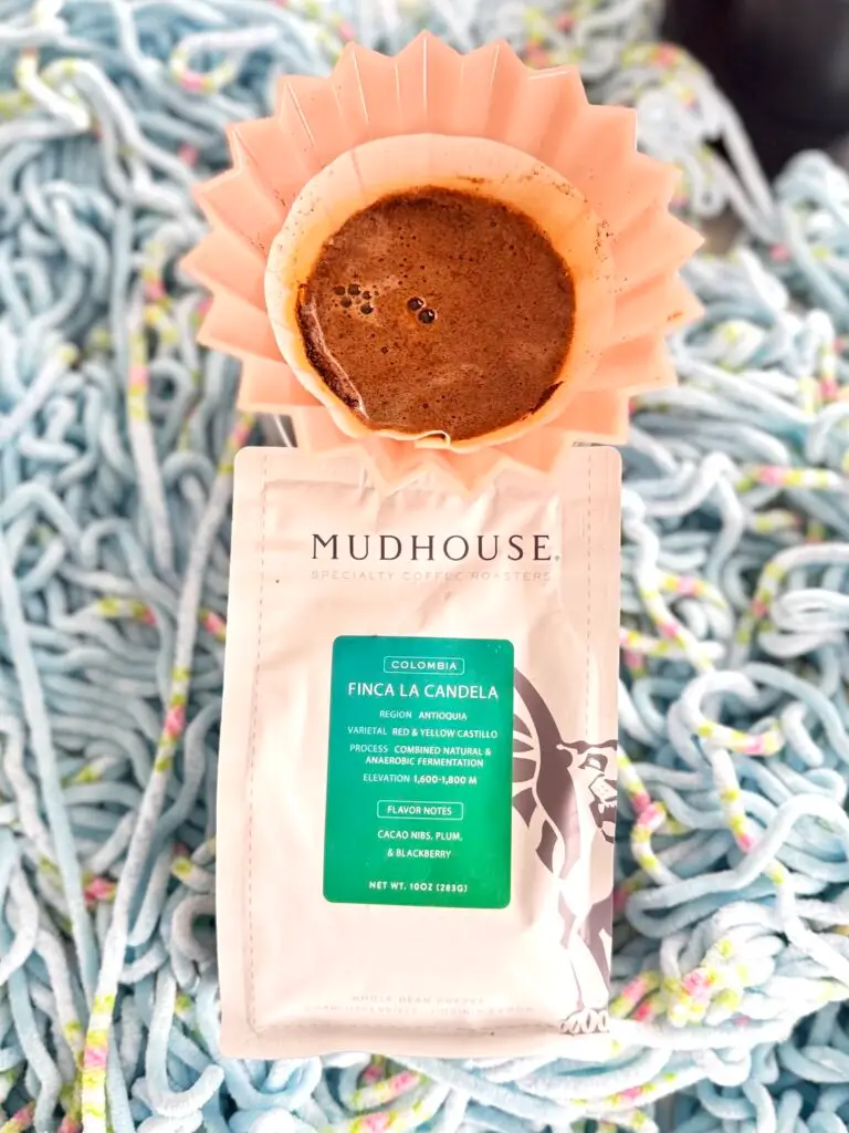 A pink coffee filter with fresh coffee sits atop a Mudhouse coffee bag. The background is a soft, pastel speckled unraveled yarn, creating a cozy atmosphere.
