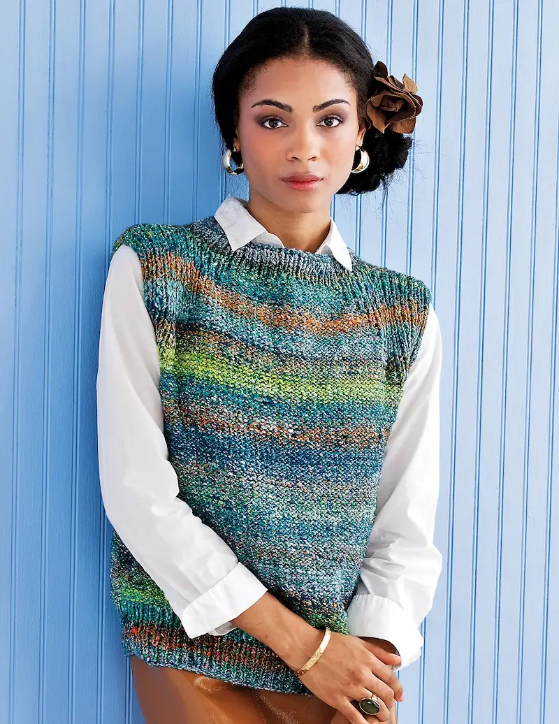 A Black woman with dark hair up in a bun models a knitted vest in variegated shades of blue, green, and orange.