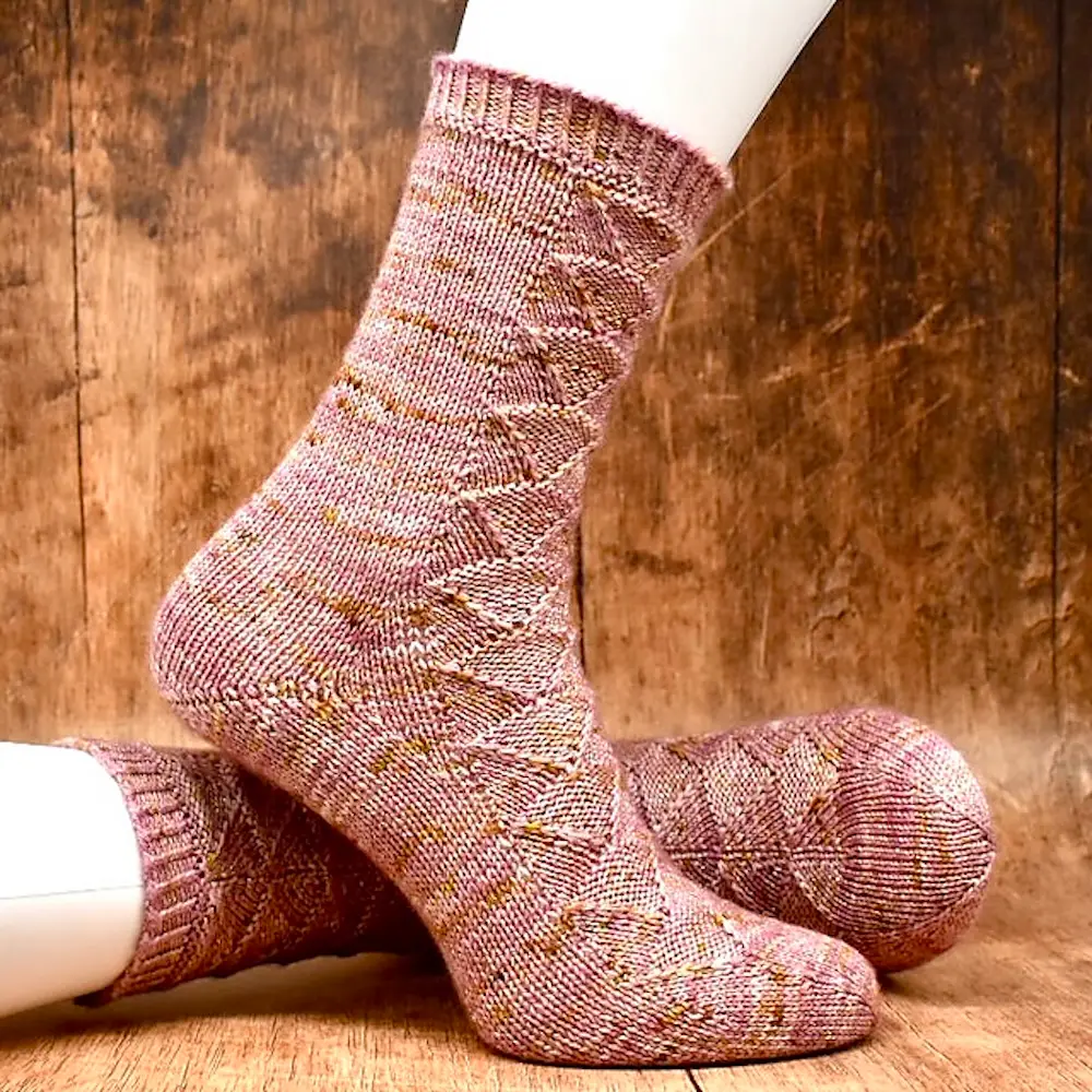 A pair of hand-knitted, crew-length socks in a variegated dusty rose and tan yarn. The socks feature an intricate textured diamond or lattice pattern throughout the leg and foot. They are displayed on white mannequin feet against a rustic, weathered wood background.