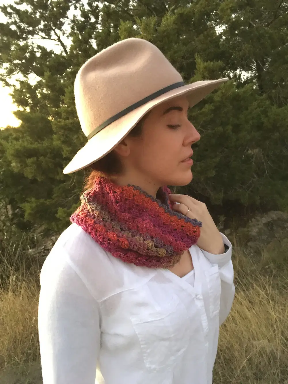 A woman in profile wears a tan wide-brimmed felt hat and a multi-colored crocheted cowl in shades of red, purple, and brown. She stands outdoors against a background of green trees and dry grass.