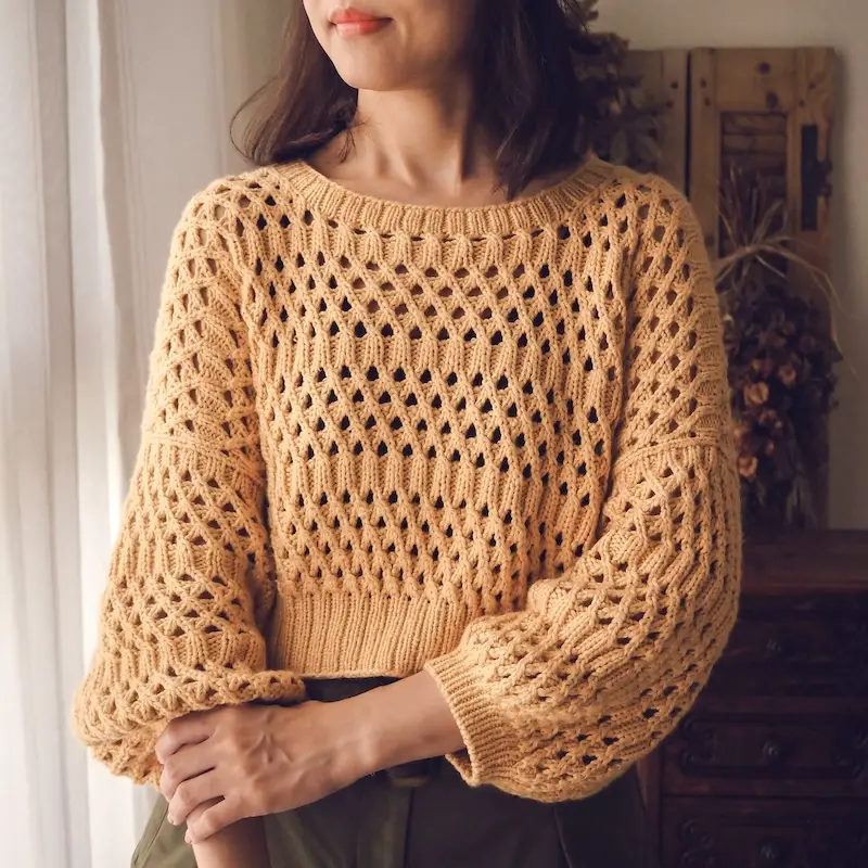 A medium-light skinned person wearing a peach-colored, hand-knitted sweater with a wide boat neck, an airy honeycomb-like eyelet pattern, and a wide ribbed hem that hits at the waist. The sleeves are long and slightly oversized, ending in simple cuffs.