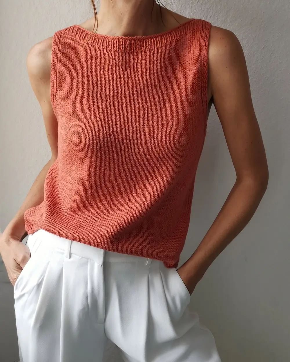 A person wearing a sleeveless, burnt-orange knit top with a high boat neckline. The top is tucked into high-waisted, pleated white trousers. The person’s hands are in their pockets, and the background is a neutral-colored wall.