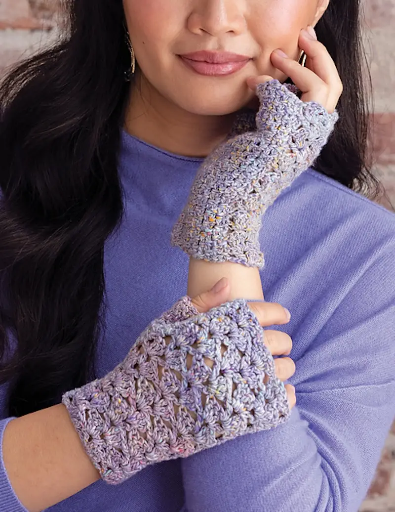 A person wearing handmade fingerless gloves made using a cabled shell crochet pattern with textured lavender yarn.