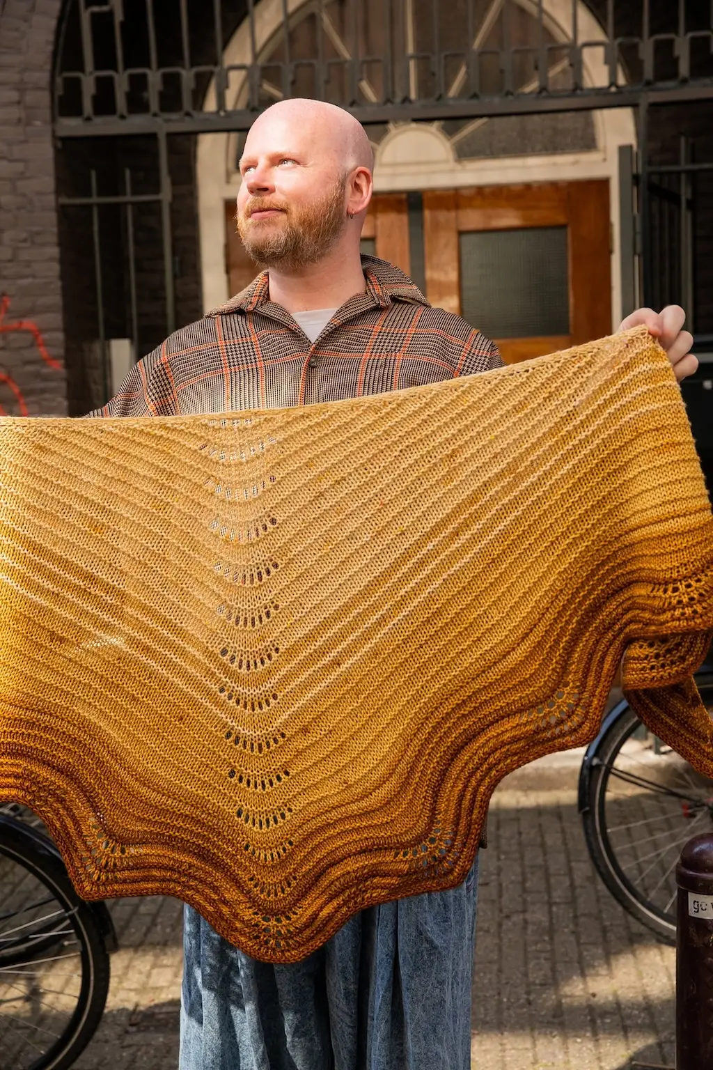 Stephen West, a white man with a reddish beard and a plaid shirt holds up a large, triangular knitted shawl in a golden-brown gradient. The shawl features horizontal garter ridges and a distinctive wavy, scalloped bottom edge.