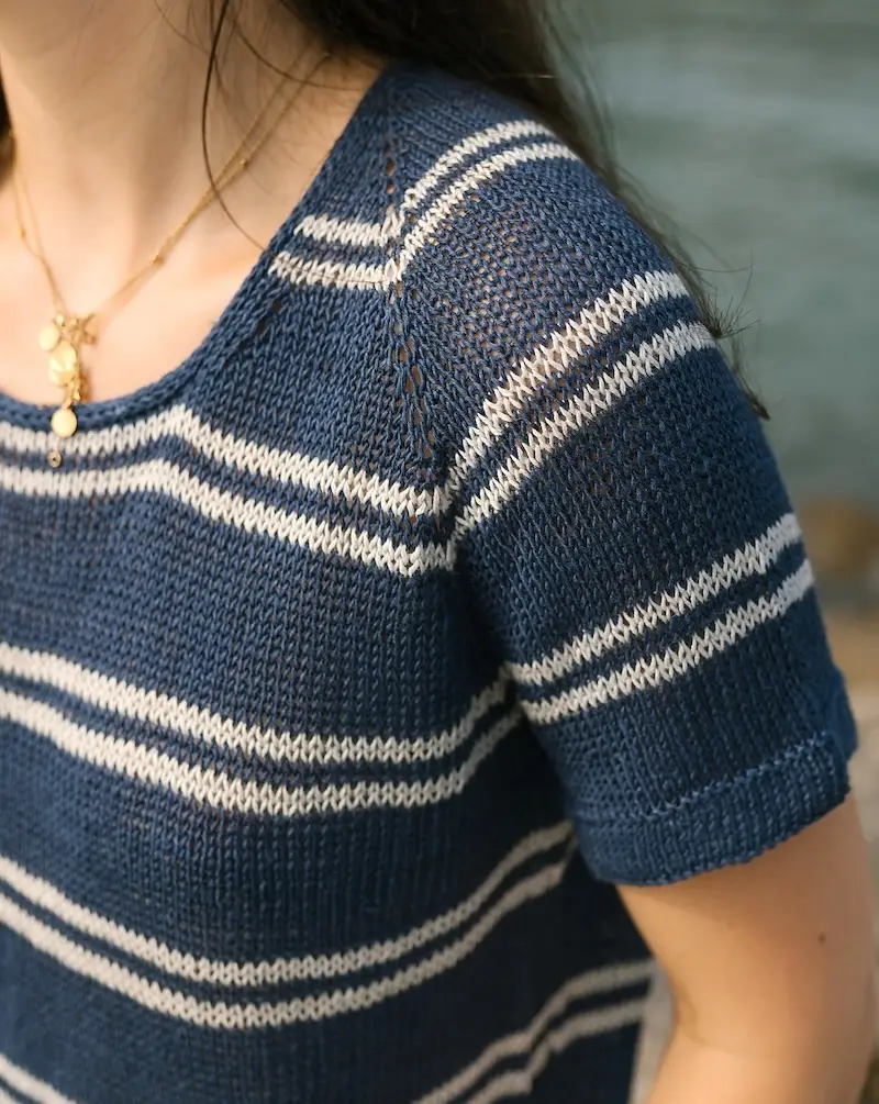 A person wearing a short-sleeved navy blue and white striped knit top. The shirt features a scoop neckline and raglan-style sleeves, with a textured, lightweight stitch pattern.