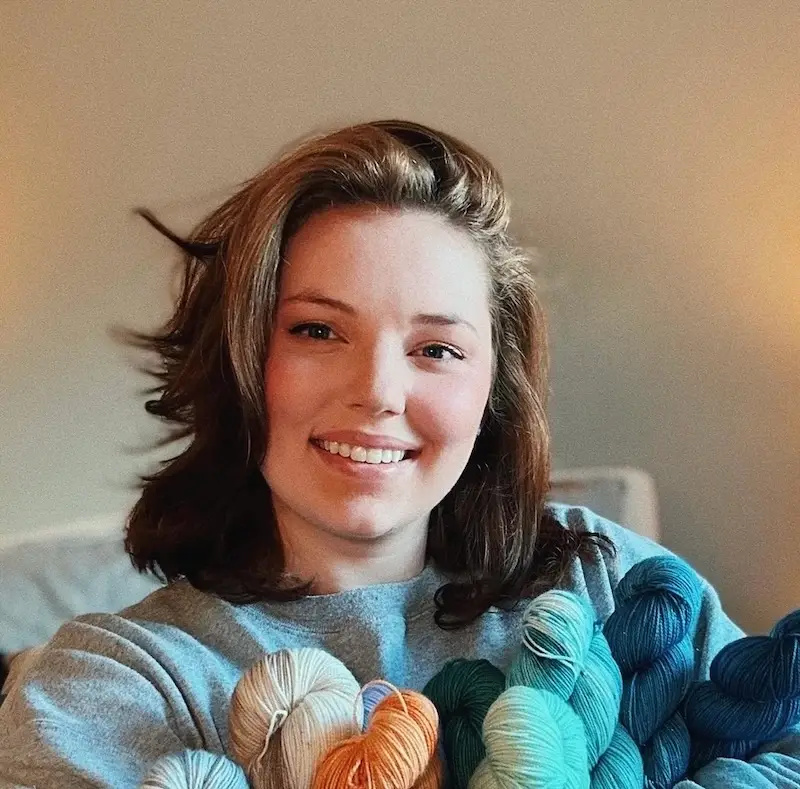 Jackson Roe, a smiling light-skinned woman with shoulder-length dark blonde hair holds an armful of colorful hand-dyed yarn skeins.